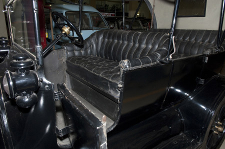 Closeup of the front driver’s seat of the car in the first image. The seat is made of black leather with a button tufted stitch pattern, and signs of wear on the armrests.